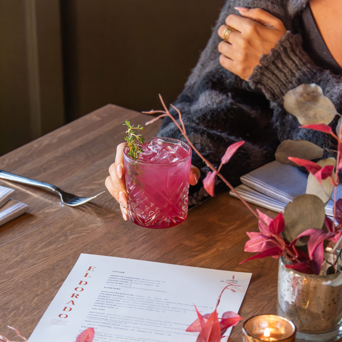 A person holding a pink cocktail with ice and a sprig of herbs at a wooden table set with a menu, foliage, and cutlery.